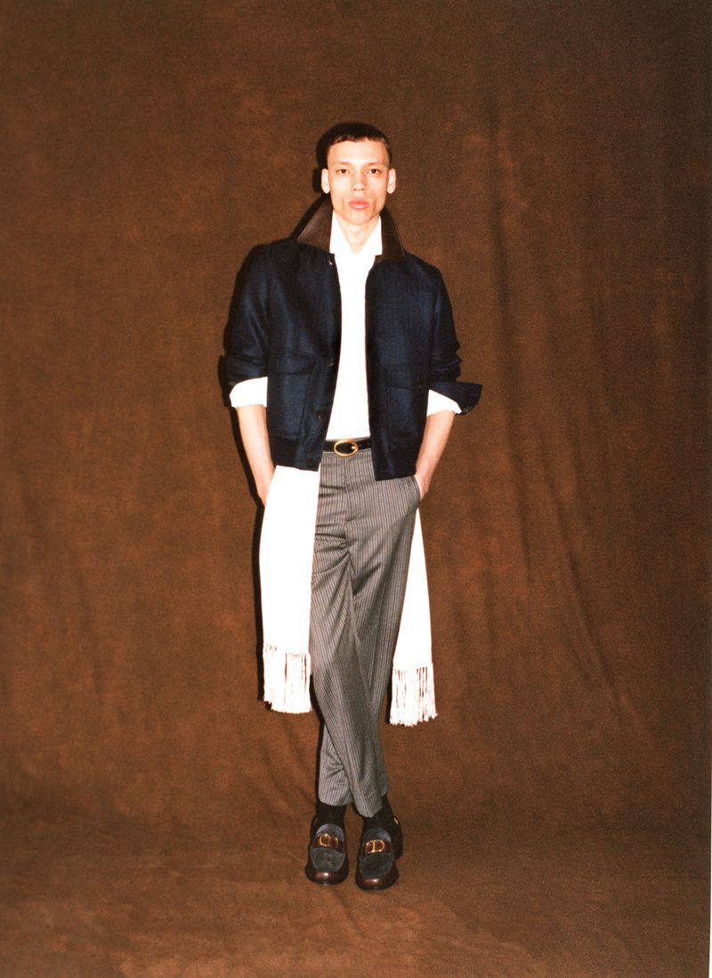 Model Jacoby Sorapuru posing in a dark cropped jacket, long white fringed shirt, and grey tailored trousers against a brown backdrop.