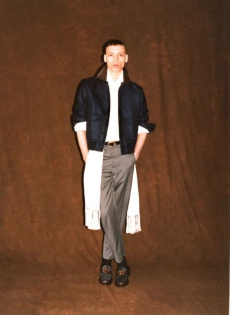 Model Jacoby Sorapuru posing in a dark cropped jacket, long white fringed shirt, and grey tailored trousers against a brown backdrop.