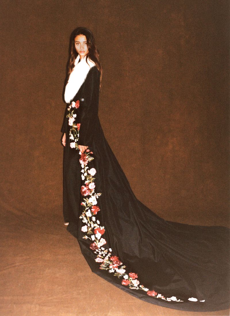 Model Jacoby Sorapuru in a long black floral-embroidered gown with a train and white fur collar against a brown backdrop.