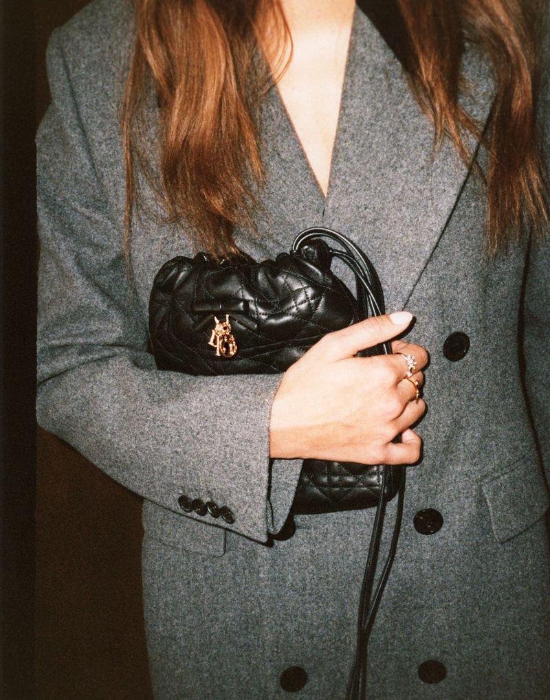 Model wearing a grey wool double-breasted coat while holding a black quilted leather mini pouch bag with gold hardware.