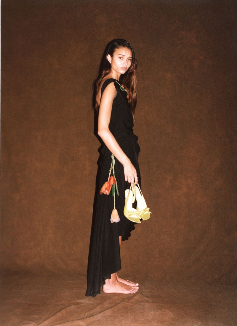 Model Jacoby Sorapuru posing barefoot in an asymmetrical black dress with floral accents against a brown backdrop for HERO USA.