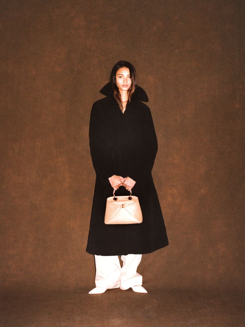 Jacoby Sorapuru in a long black high-collar coat holding a beige handbag against a warm brown textured backdrop.