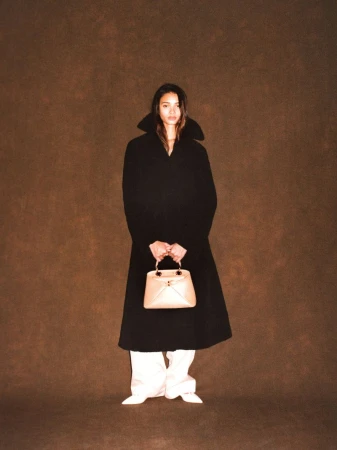 Jacoby Sorapuru in a long black high-collar coat holding a beige handbag against a warm brown textured backdrop.