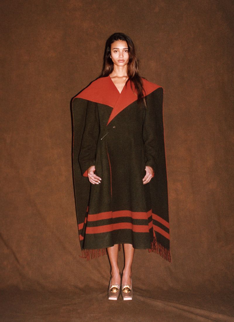 Model wearing a dark olive and rust-red wool cape coat with fringe trim against a warm brown textured backdrop.