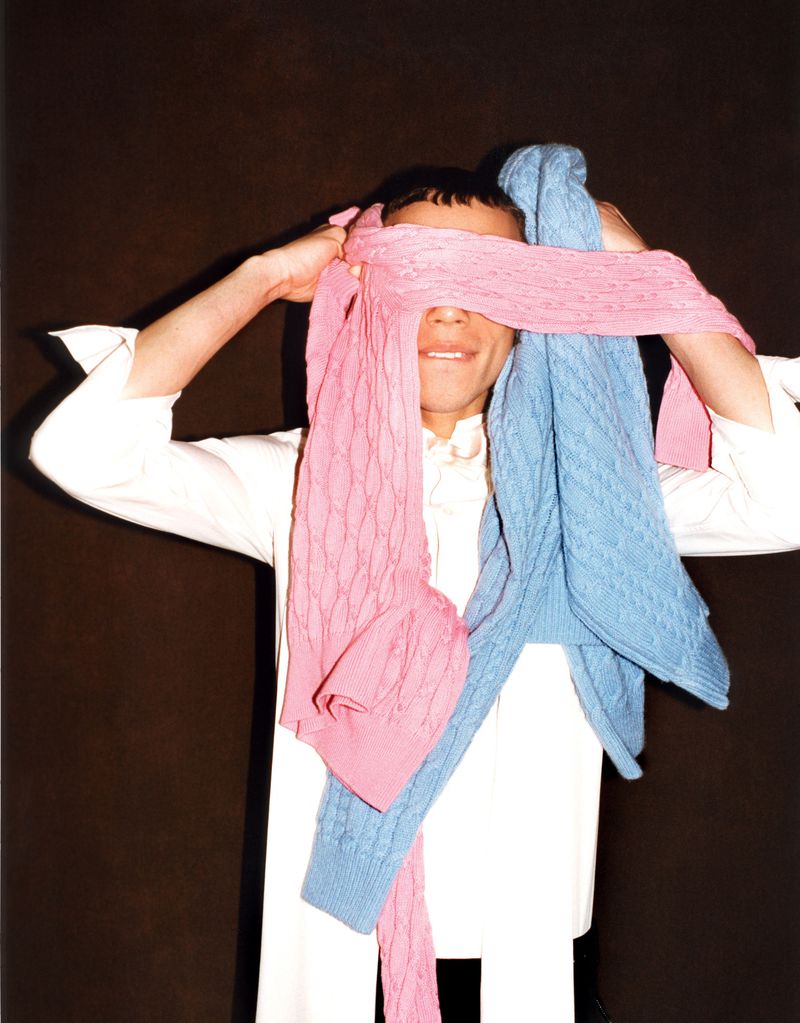 Model with eyes covered by draped pink and blue cable knit sweaters wearing a white collared shirt against a dark backdrop.