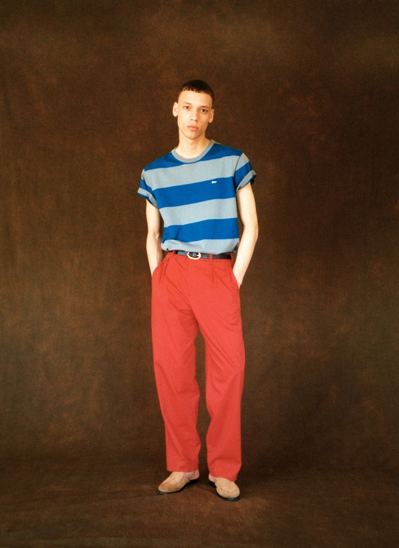 Model Jacoby Sorapuru poses in a blue striped t-shirt and high-waisted red trousers against a mottled brown studio backdrop.
