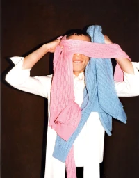 Model with eyes covered by draped pink and blue cable knit sweaters wearing a white collared shirt against a dark backdrop.