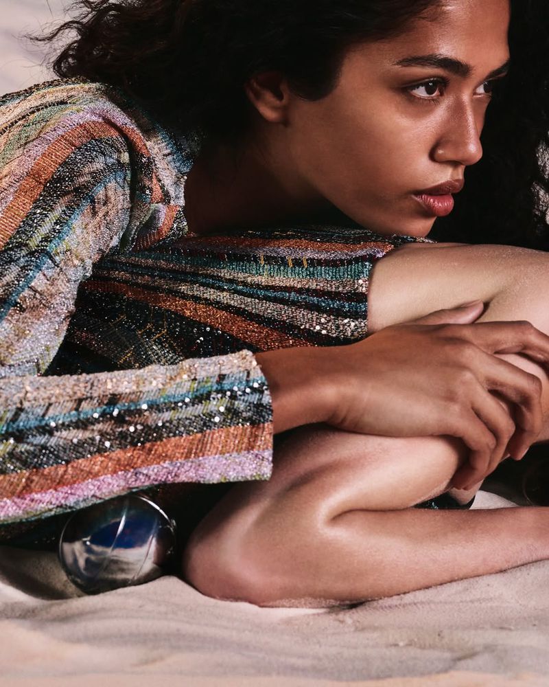 Model Jessie Aina seated on sand wearing a multi-colored metallic striped knit top with her knees drawn in close.