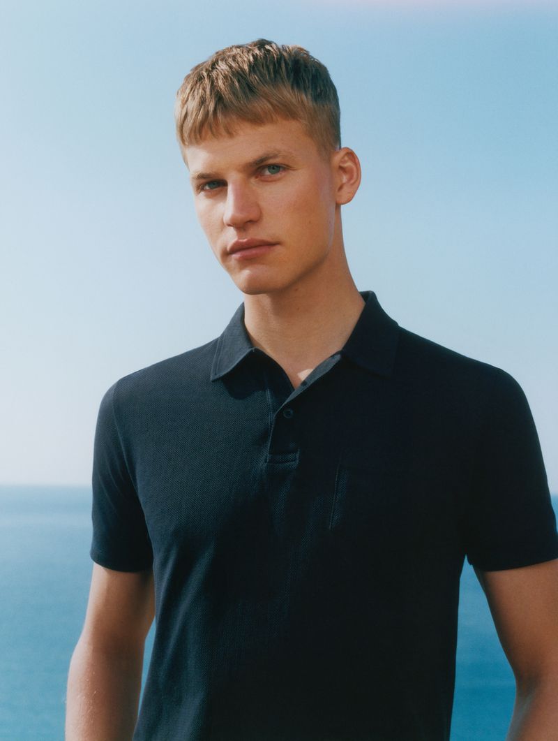 Man with cropped blonde hair wearing a black short-sleeved polo shirt with an open ocean and clear sky background.