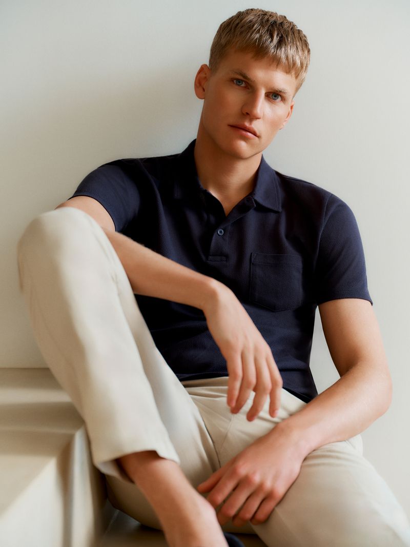 Model Timo Pan wearing a navy blue polo shirt and beige trousers seated against a neutral studio backdrop for Sunspel.