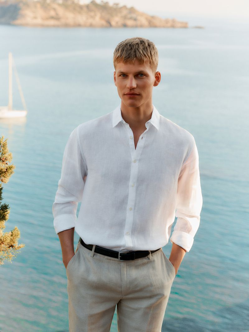 Model wearing a white linen button-up shirt and beige trousers standing against a calm coastal sea background.