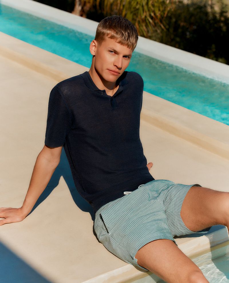 Model Timo Pan sitting poolside in a navy knit polo shirt and green striped swim shorts in direct sunlight.