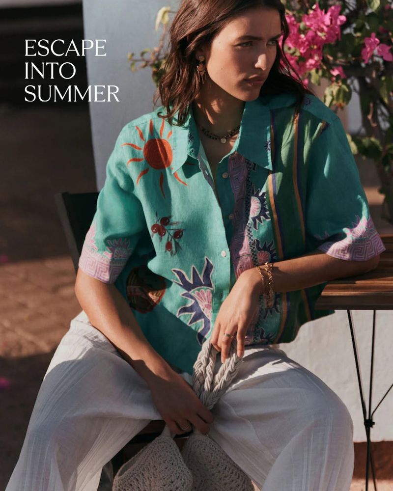 Model Adele Aldighieri wearing a teal printed shirt and white trousers seated outdoors near bougainvillea flowers.