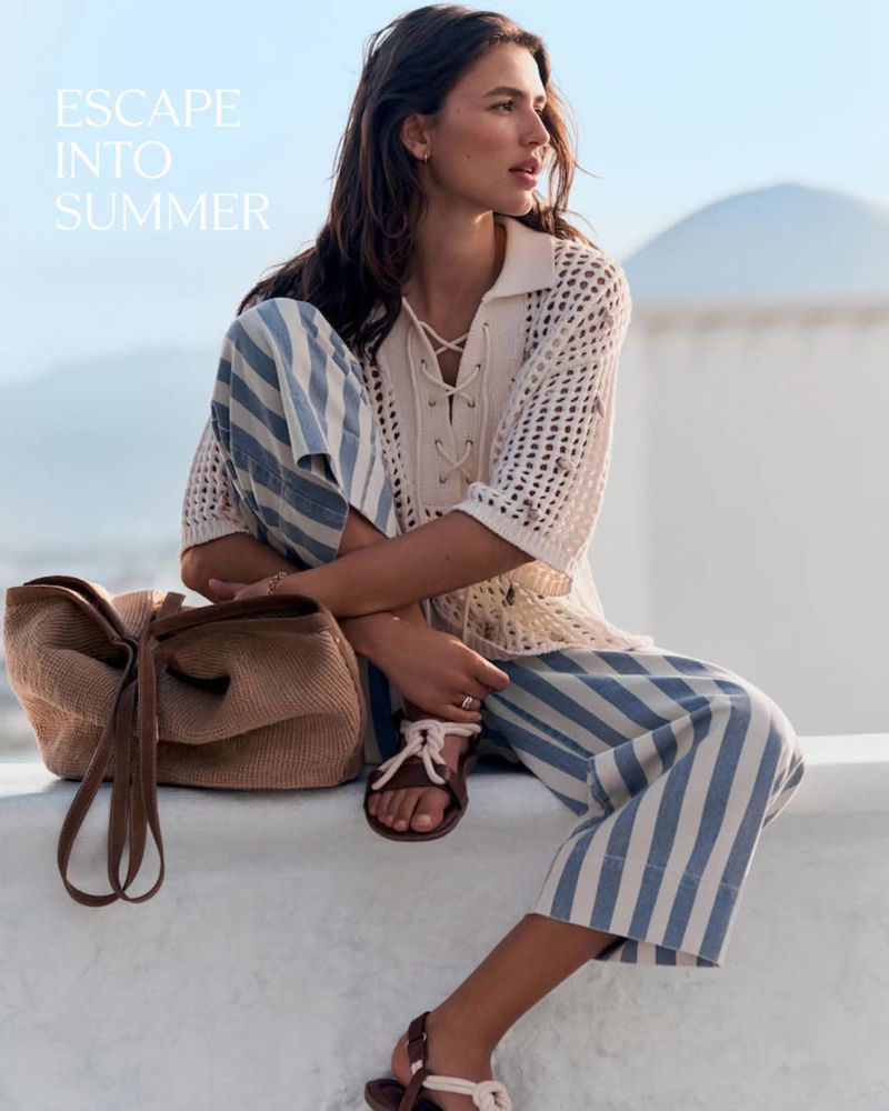 Model Adele Aldighieri wearing a crochet polo and striped trousers on a sunlit whitewashed terrace.