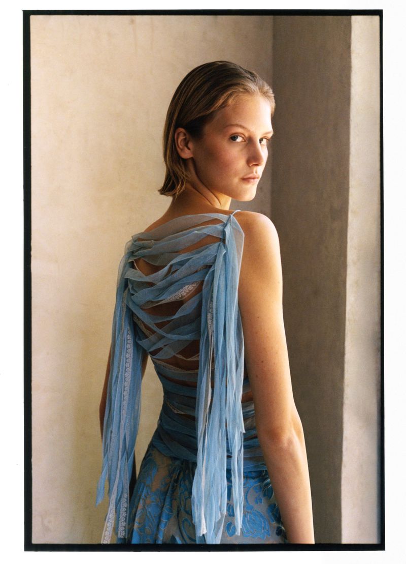 Model wearing a blue tulle dress looking over her shoulder indoors against a neutral wall
