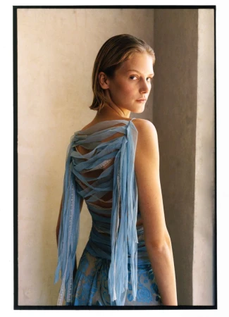 Model wearing a blue tulle dress looking over her shoulder indoors against a neutral wall