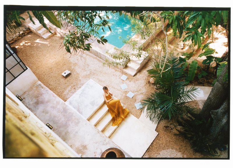 Model Skye Blendermann sitting on stone courtyard steps wearing a flowing yellow gown beside a swimming pool.