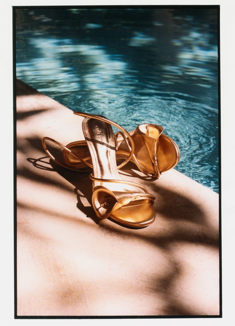 Pair of metallic gold high heel sandals resting on a stone ledge beside a sunlit blue swimming pool.