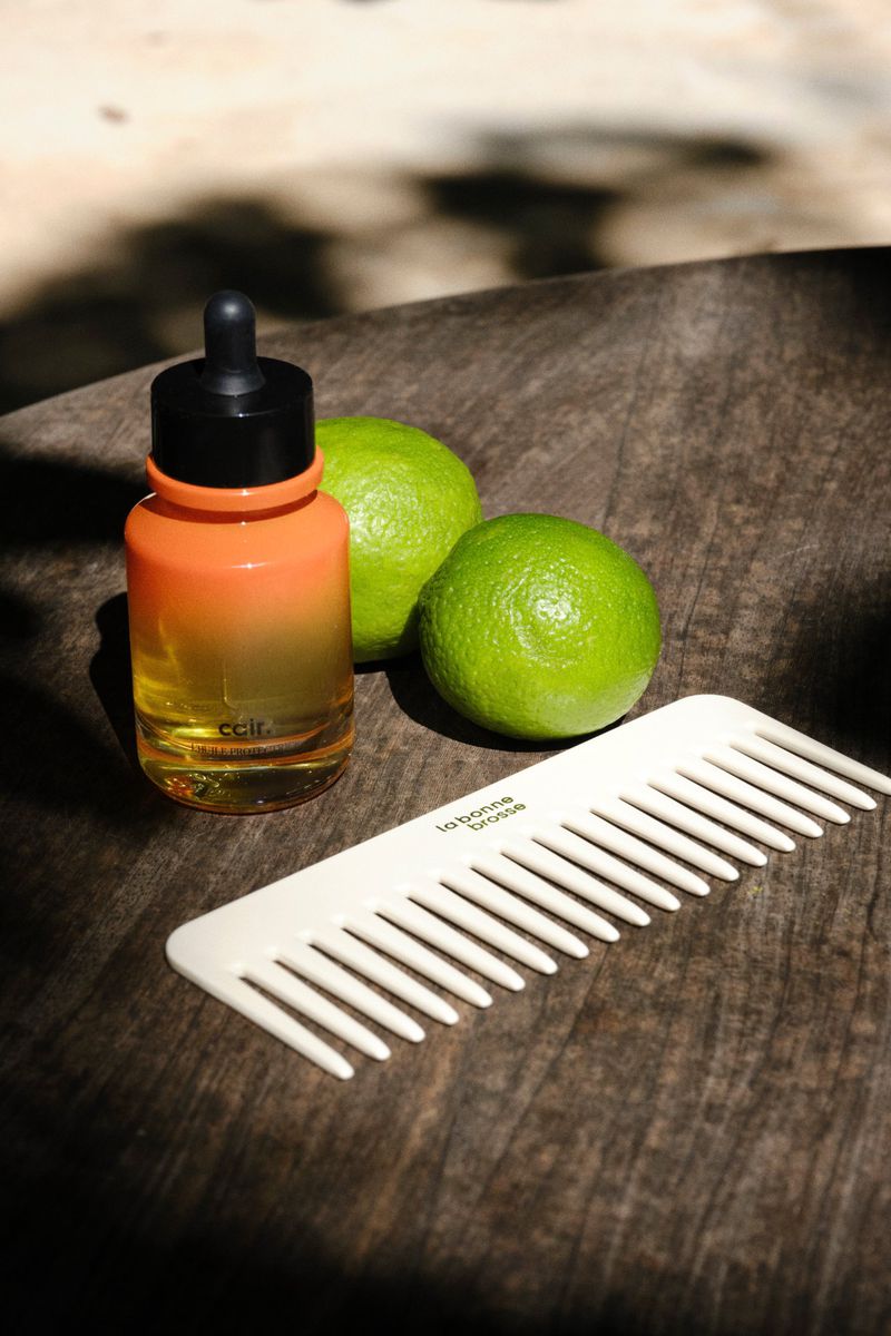 Dropper bottle of oil, two green limes, and a wide-tooth comb arranged on a dark wooden table in dappled sunlight.
