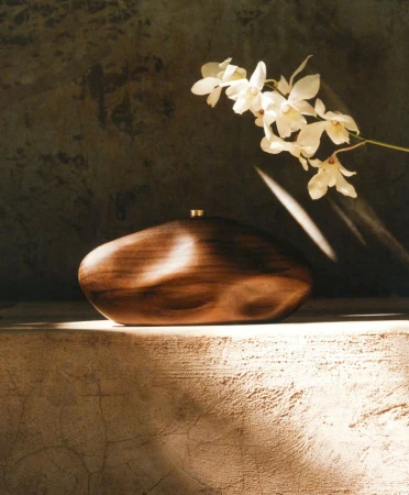Sculptural polished wooden clutch with gold clasp resting on a textured concrete ledge beside white orchid blossoms.