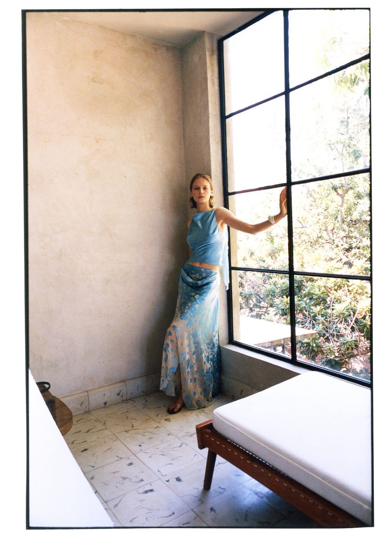 Skye Blendermann in a light blue top and patterned maxi skirt posing beside a large grid window for Moda Operandi.