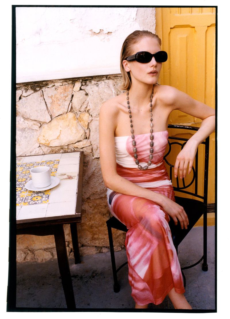 Model Skye Blendermann wearing dark sunglasses and a strapless printed dress seated at an outdoor cafe table.