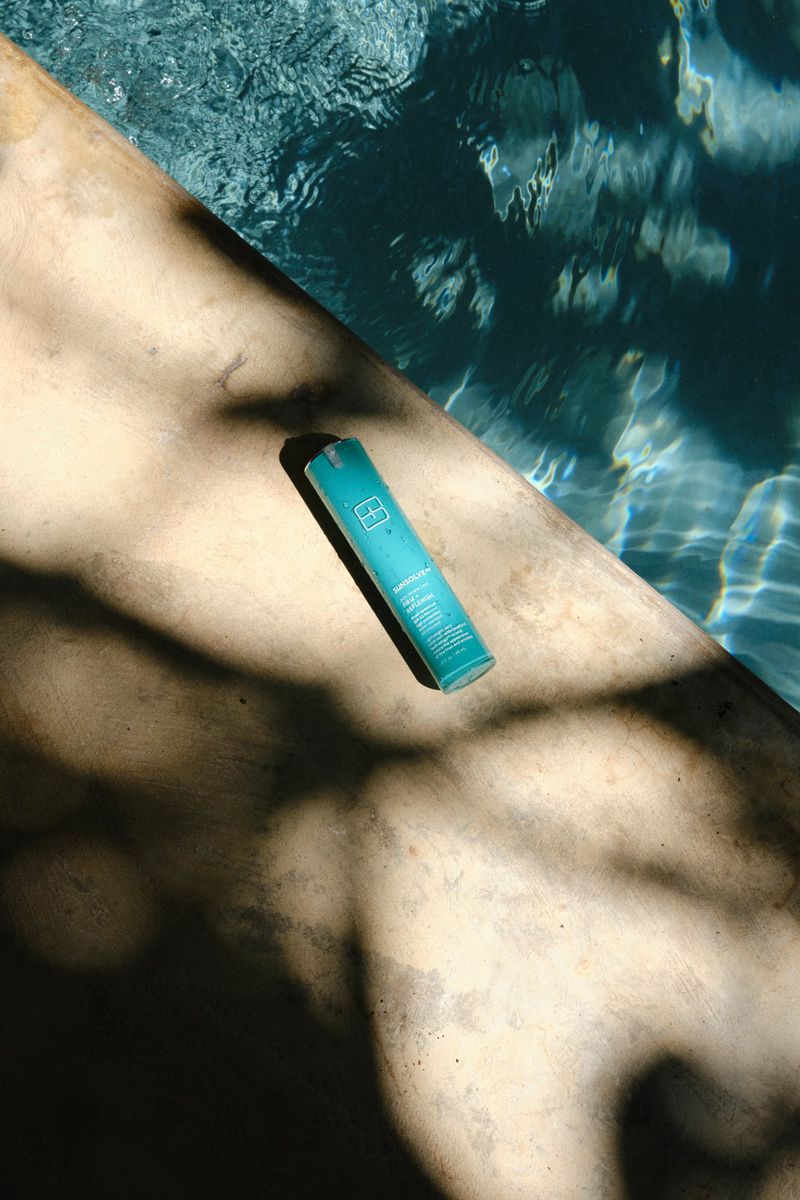 A teal skincare bottle rests on a concrete poolside ledge cast in dappled tree shadows next to clear blue pool water.