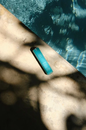 A teal skincare bottle rests on a concrete poolside ledge cast in dappled tree shadows next to clear blue pool water.