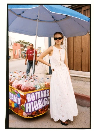 Model Skye Blendermann in a white eyelet dress and sunglasses posing next to a snack cart under an umbrella on a street.