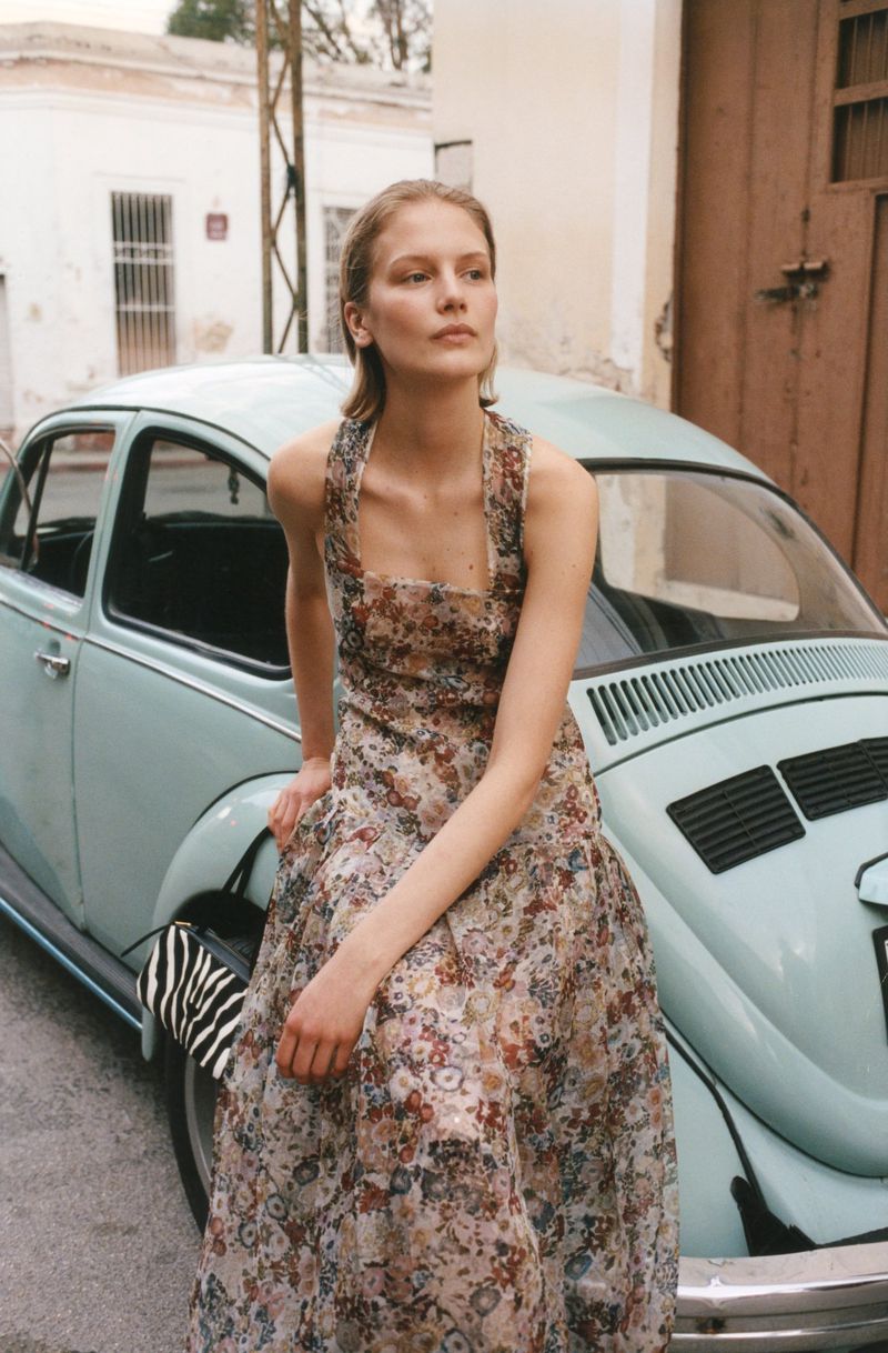 Model Skye Blendermann wearing a floral maxi dress seated on the hood of a vintage light blue car along a street.