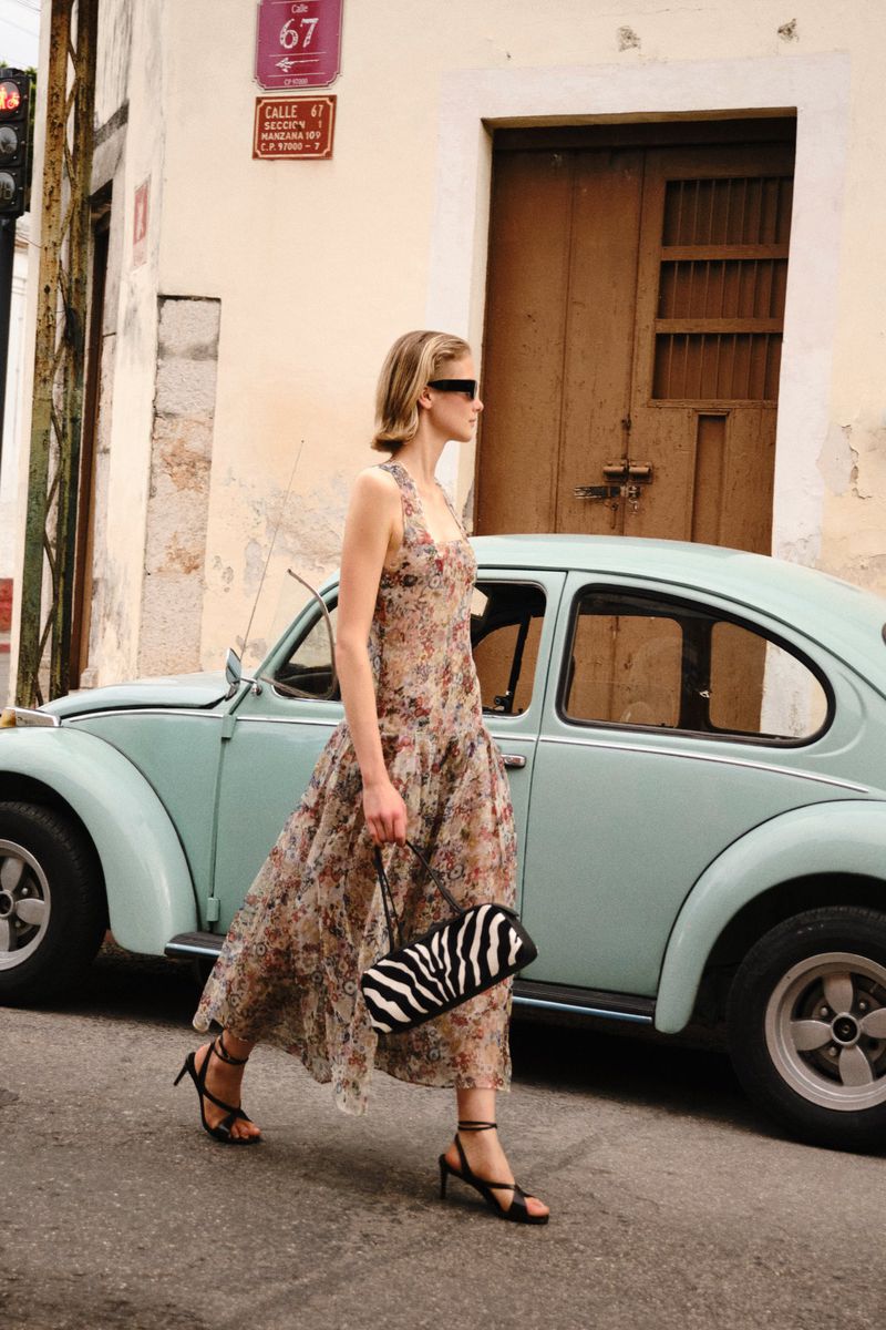 Model Skye Blendermann walking down a street in a sheer floral dress and heels past a vintage light blue car.