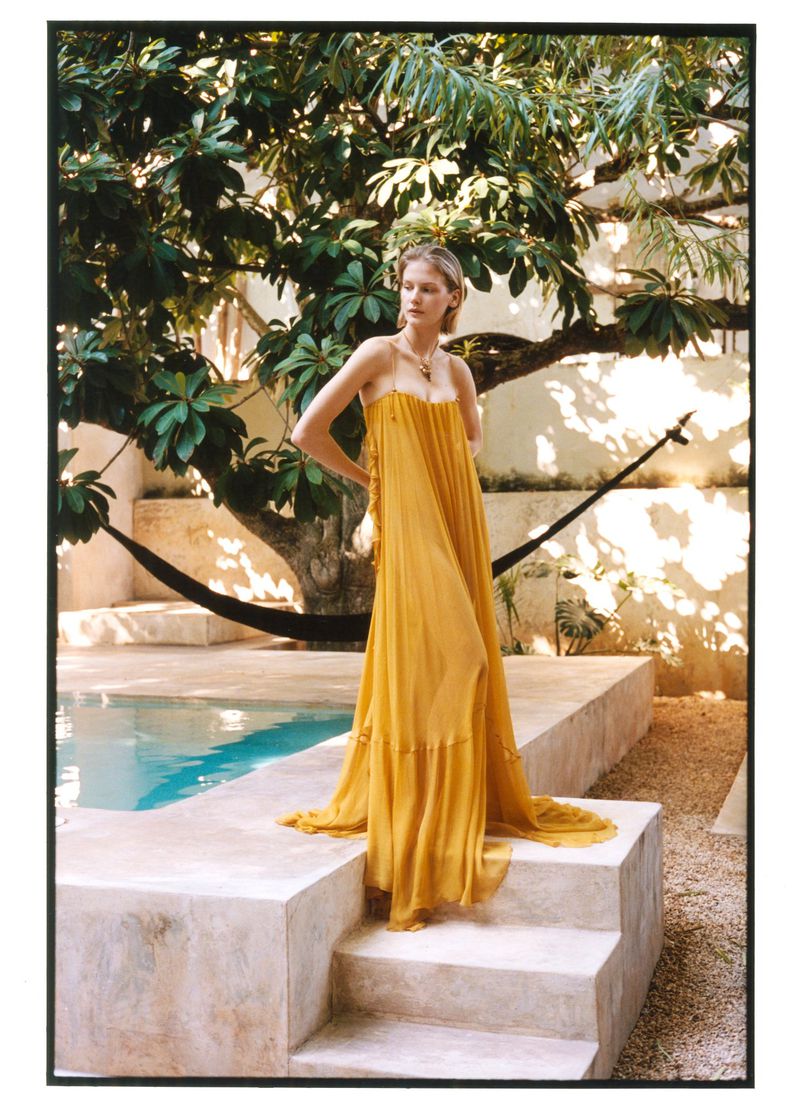 Model wearing a flowing yellow floor-length dress standing on stone steps by an outdoor pool surrounded by lush foliage.