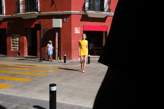 Model in a bright yellow mini dress walking across a sunlit street crosswalk in front of a red building facade.