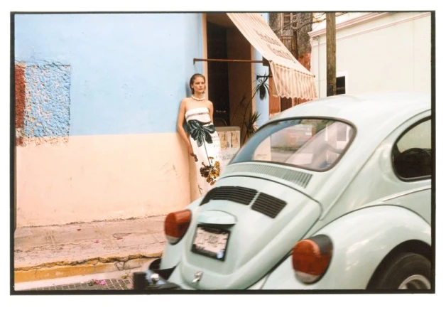 Model Skye Blendermann in a printed strapless dress leans on a pastel wall beside a vintage light blue car.