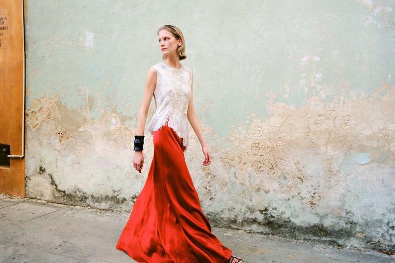 Model Skye Blendermann walking along a textured wall wearing a sleeveless white lace top and long red satin skirt.