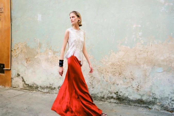 Model Skye Blendermann walking along a textured wall wearing a sleeveless white lace top and long red satin skirt.