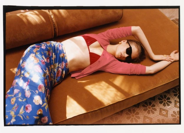 Model Skye Blendermann reclining on a velvet daybed wearing a floral blue skirt, pink cardigan, and dark sunglasses.