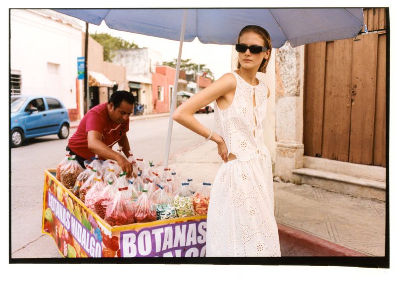 Model Skye Blendermann wearing a white eyelet dress and sunglasses next to a colorful snack cart on an outdoor street.