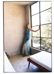 Skye Blendermann in a light blue top and patterned maxi skirt posing beside a large grid window for Moda Operandi.