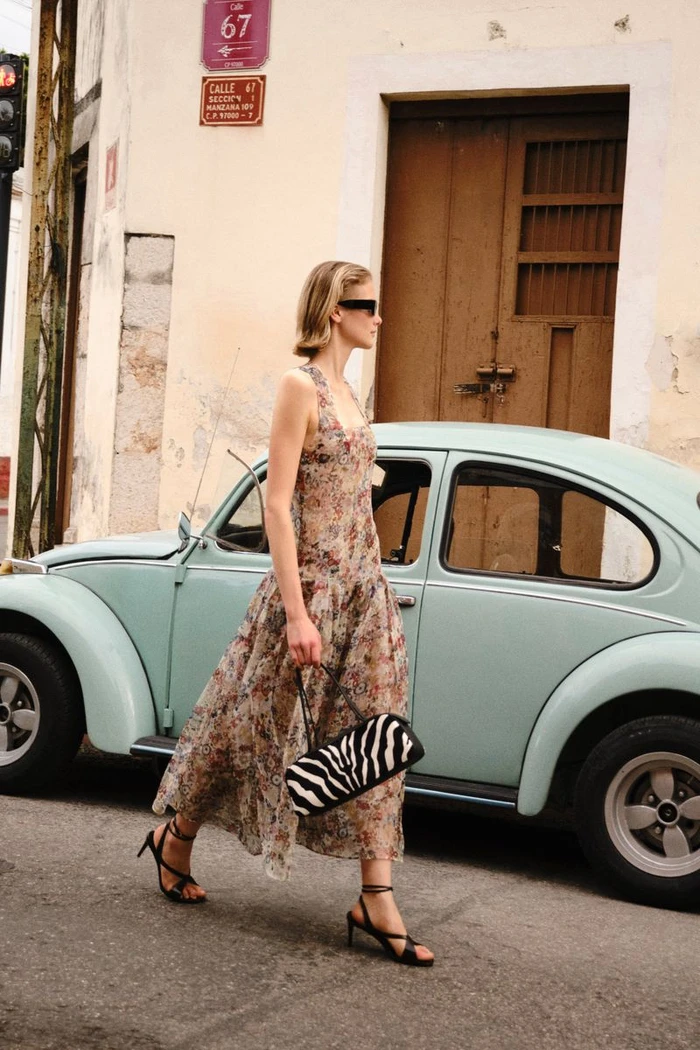 Model Skye Blendermann walking down a street in a sheer floral dress and heels past a vintage light blue car.