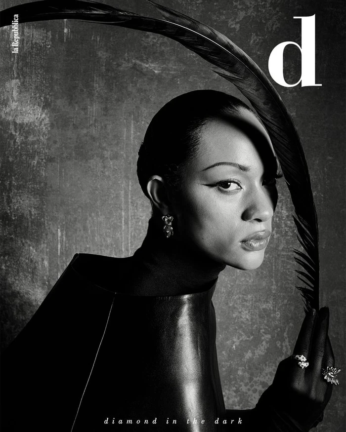 Selena Forrest holding a curved black feather above her head wearing black leather and fine jewelry against a textured studio backdrop.