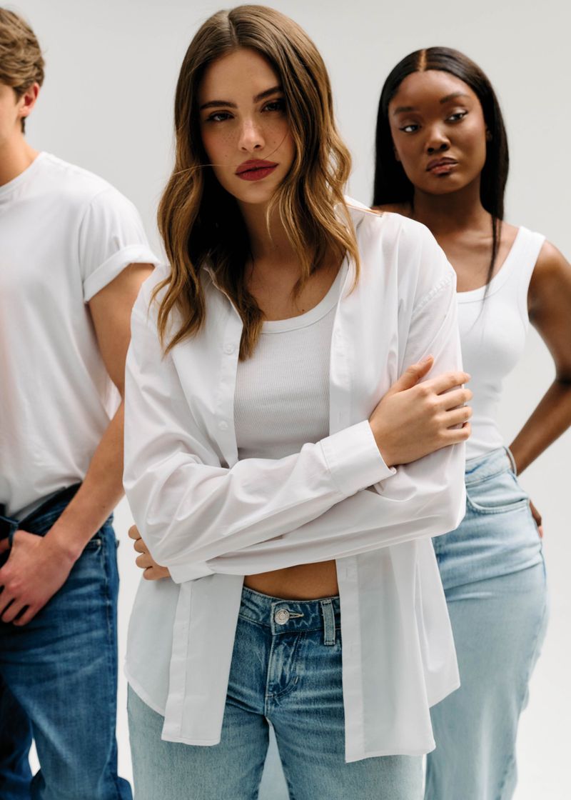Woman in an unbuttoned white shirt and blue jeans posing between two models against a seamless white backdrop.