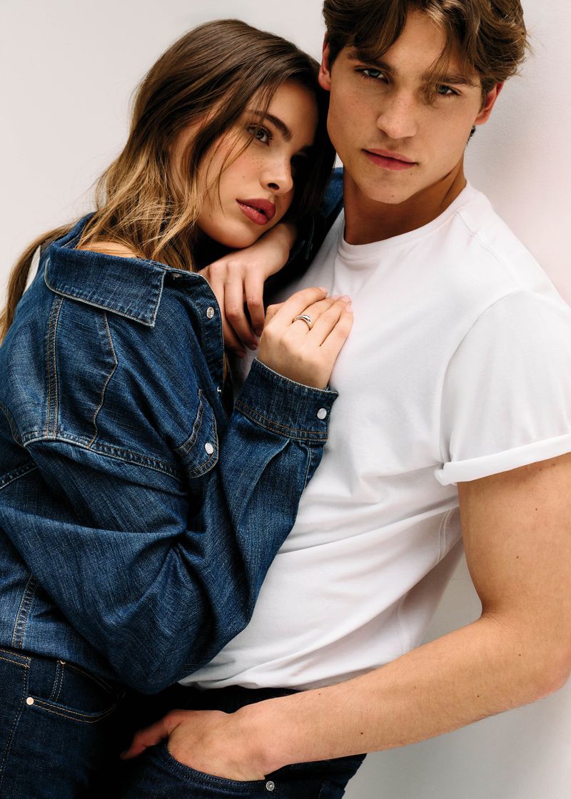 A woman in a denim jacket rests her head and hand on a man wearing a white rolled-sleeve t-shirt against a white background.