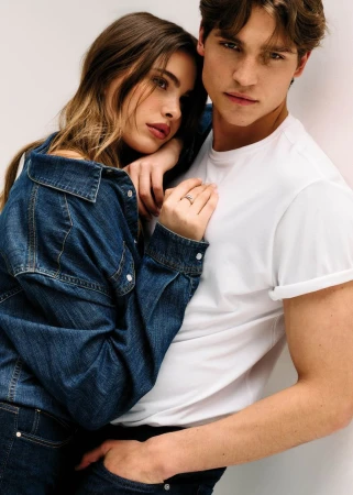 A woman in a denim jacket rests her head and hand on a man wearing a white rolled-sleeve t-shirt against a white background.