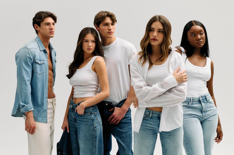 Five fashion models posing together wearing white tops and classic blue denim jeans against a white seamless background.
