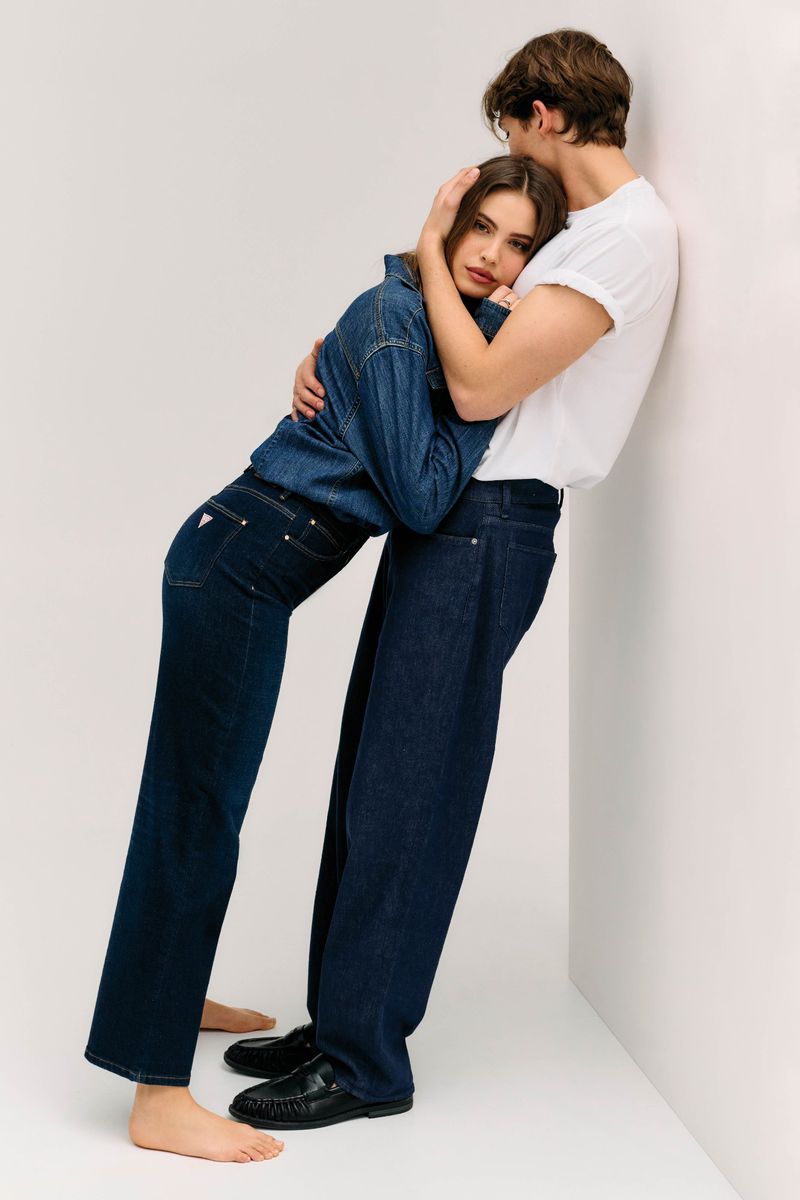 A man in a white t-shirt and woman in a denim jacket embracing, leaning against a white studio wall