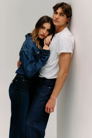 A woman in a dark denim jacket and jeans leans against a man wearing a white t-shirt and denim trousers by a white studio wall.