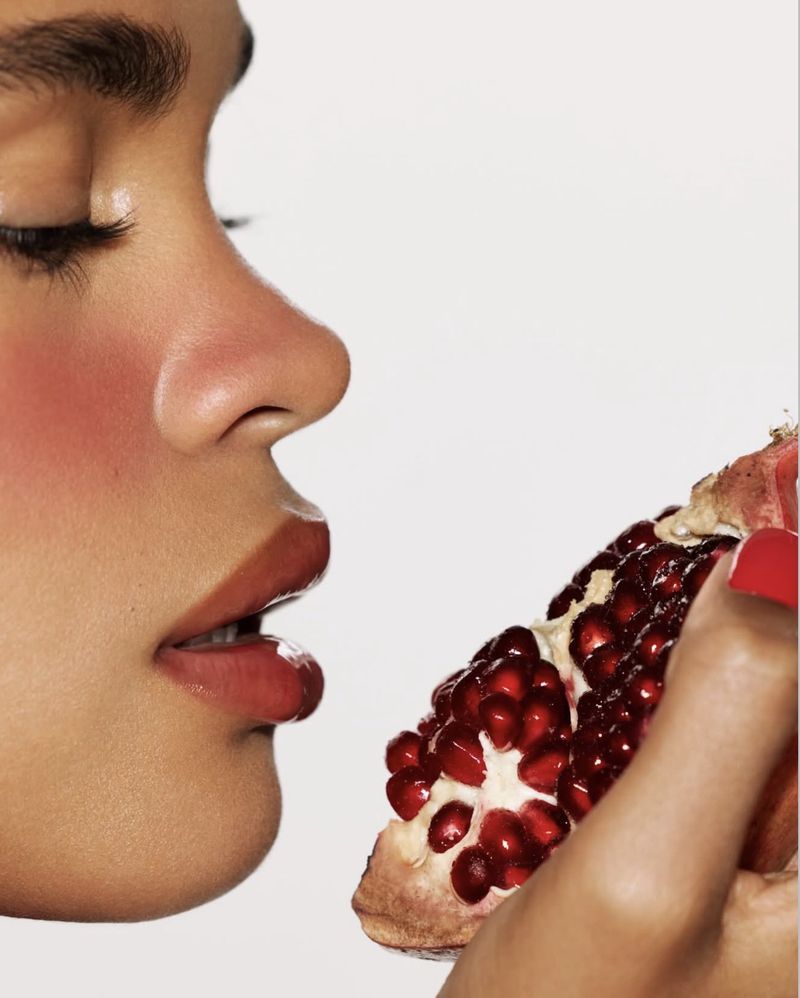 Close-up profile of a woman with glossy lips holding an open pomegranate with visible red seeds against a white studio background.