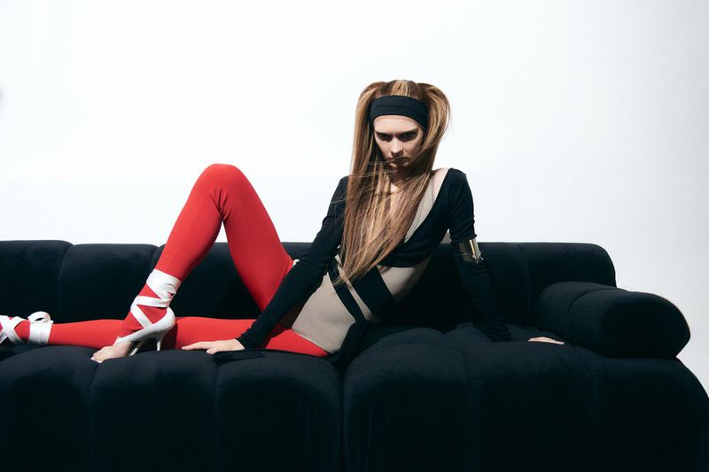 A woman with long hair poses on a black couch in a black and beige bodysuit with red leggings and wrapped heels in a photo studio.