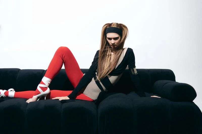A woman with long hair poses on a black couch in a black and beige bodysuit with red leggings and wrapped heels in a photo studio.